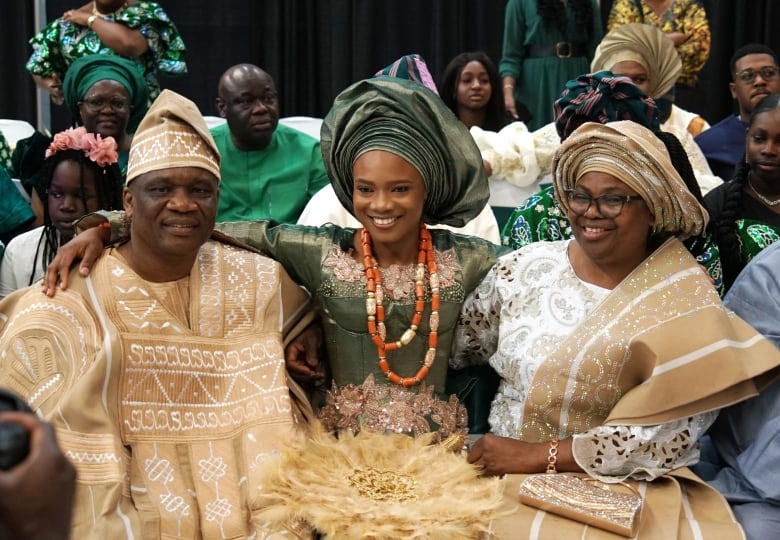 A young woman in traditional green Nigerian dress embraces an older man and woman in traditional cream robes.