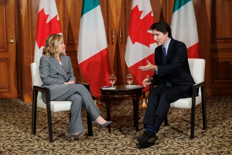 People sitting in chairs speak to each other in front of a line of Canadian and Italian flags.