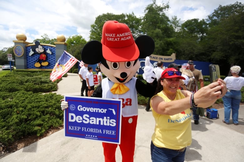 A person wears a costume closely resembling Mickey Mouse, with an oversized red hat that says Make American Great Again, holds a sign that says "DeSantis Keep Florida Free" A woman in a T-shirt takes a selfie with the mouse on her smartphone.