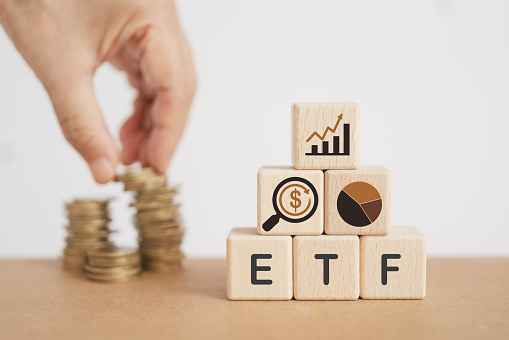 wooden cube with ETF text , Exchange Traded Fund and blurred hand with stack of coins. for business finance conceptual