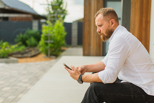 Young blonde man working at home. Back yard of house. Businessman with mobile phone