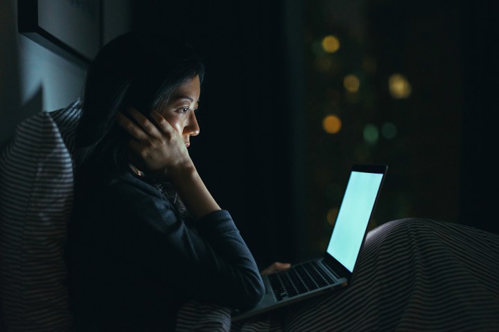 Young woman in deep thought while using laptop on bed at night.