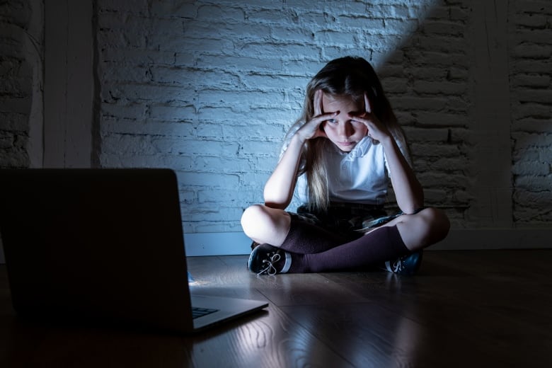 Looking distraught, a young girl sits crosslegged on the floor in a darkened room with her head in her hands, peering at an opened laptop facing her.