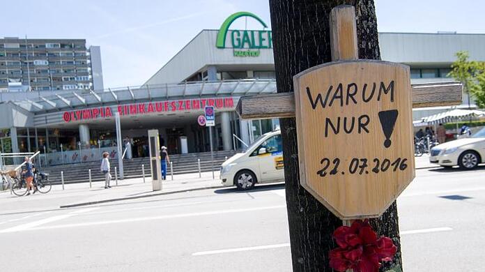 Ein Holzkreuz mit einem Schild und der Aufschrift "Warum nur! - 22.07.2016" hängt vor dem Eingang zum Olympia-Einkaufszentrum (OEZ).