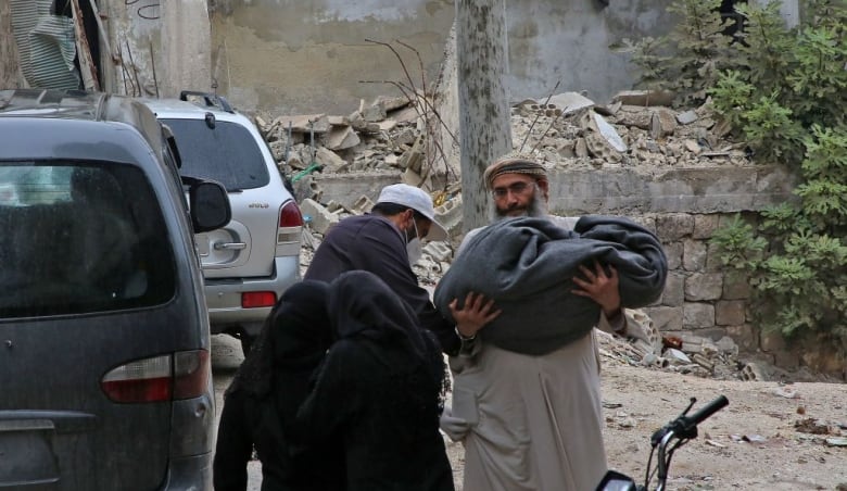 A man carries a body in a blanket past the rubbled of a destroyed building.