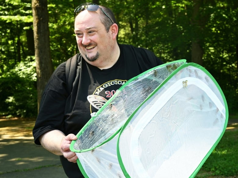 A smiling man stands outside carrying a rectangular netted enclosure full of big winged insects.