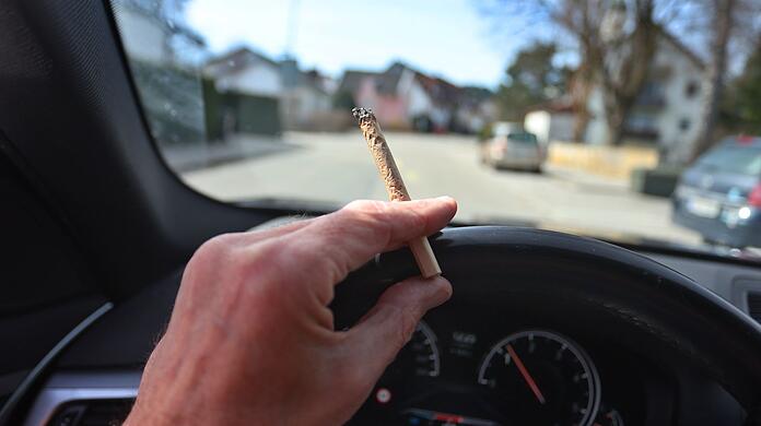 Ein Mann sitzt mit einem Joint zwischen den Fingern am Steuer eines Autos.