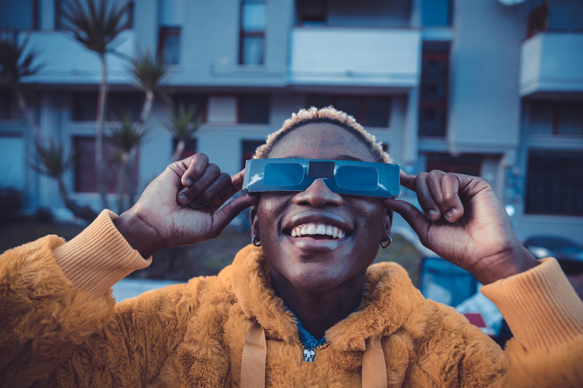 A person in a yellow fuzzy sweatshirt wearing solar eclipse glasses outside