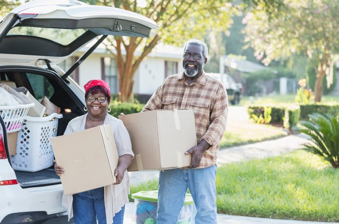 Two people holding moves boxes next to a car with an open trunk.