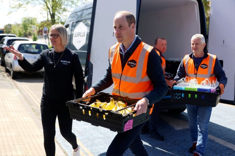 A person carries a box of bananas with other people in the background, along with a van.