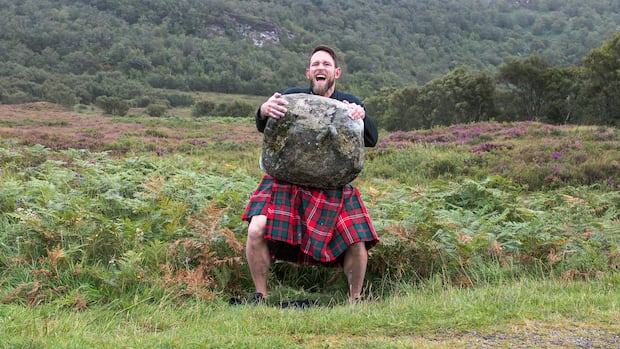 Dale Nisbet lifts a stone, wearing a traditional kilt, in front of grassy hills on an overcast day.