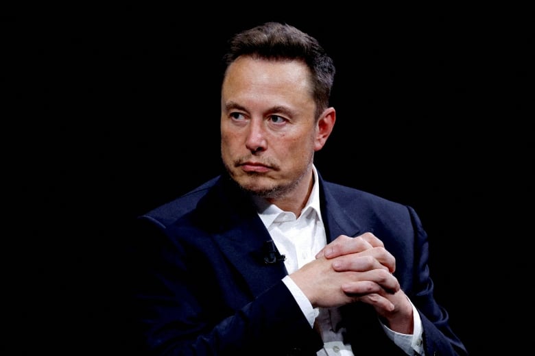 A man wearing a blazer and white collared shirt is shown with hands clasped, appearing to listen to someone off camera.