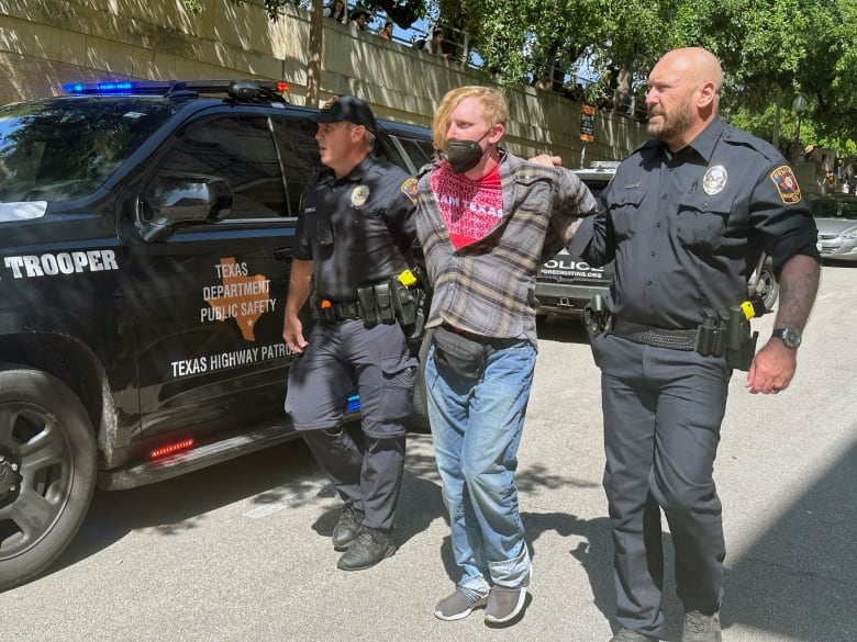 A young man with a face covering is led by police officers on either side of him in a daytime photo outdoors.