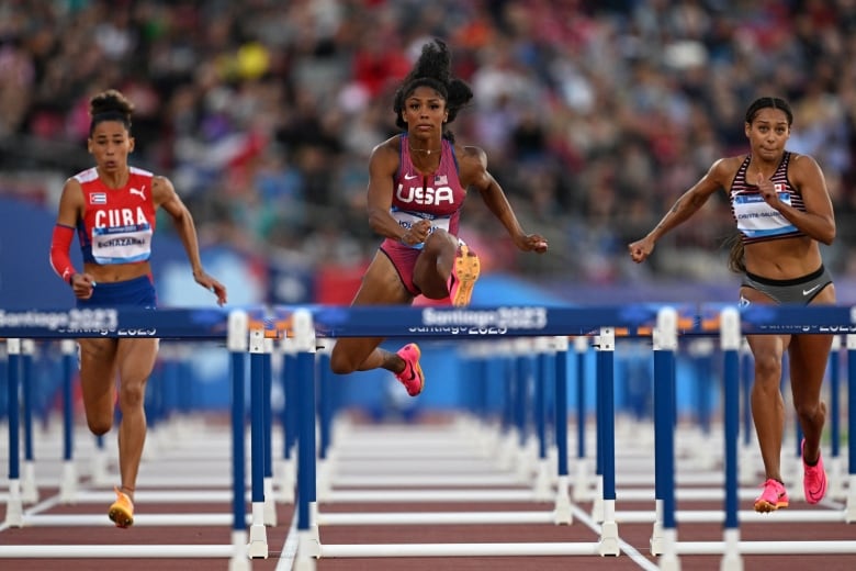 Three female athletes hop over hurdles on a track