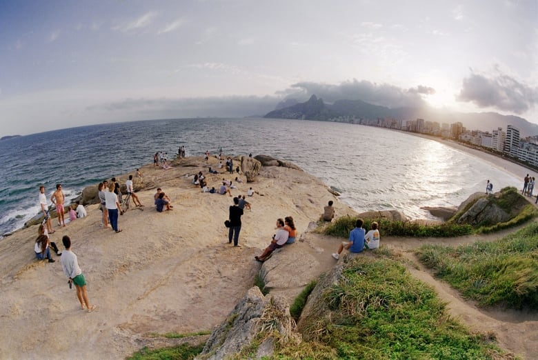 A wide angle view of people on a beach, with a city in the far background. The sun is particularly bright in the sky.