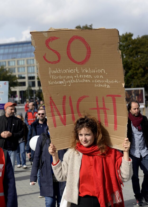 Eine Frau mit einem roten Schal trägt ein Pappschild auf den Schultern, daruf steht: 