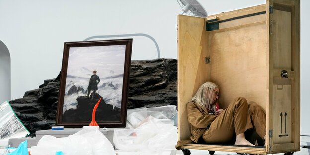 Eine Frau sitzt in einer Holzbox, daneben Caspar David Friedrichs Gemälde 