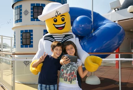 Ein Junge und ein Mädchen stehen auf einem Kreuzfahrtschiff vor einer lebensgroßen Legofigur und schießen ein Selfie