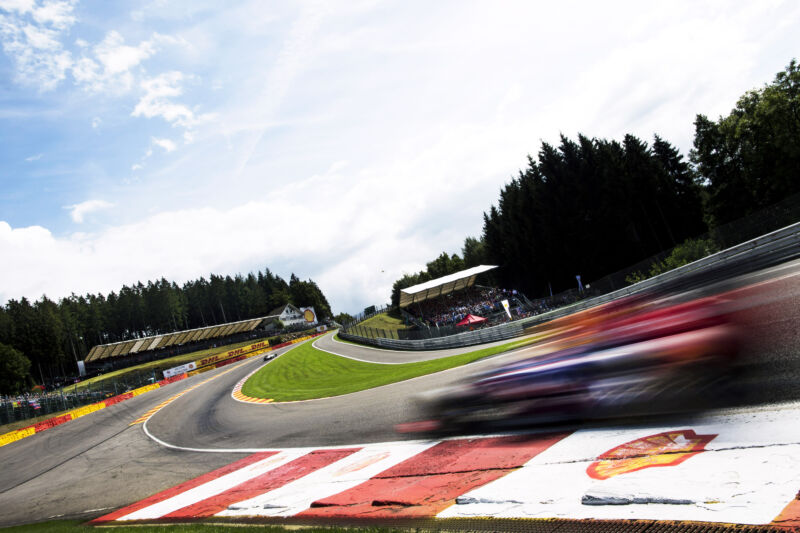 Under a cloudscape sky, and in front of trees of the Ardennes Forest, a Red Bull Racing RB10 racing car driven at speed by either German Sebastian Vettel or Australian driver Mark Webber through the Eau Rouge corner and towards the Raidillon corner following other cars while being watched by a crowd of people sitting in the grandstand during the race at the 2014 Belgian Grand Prix, Spa-Francorchamps, Belgium, on the 24th August 2014. (Photo by Darren Heath/Getty Images)