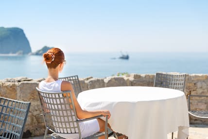 Eine Frau sitzt an einem Tisch in der Sonne, mit einem tollen Ausblick auf das Meer.