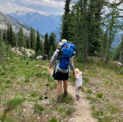 A woman holds a child's hand as they walk on a path with trees and mountains in the distance. 