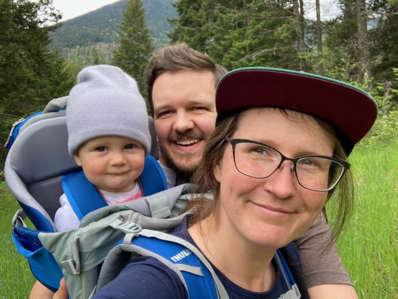 A closeup photo shows a smiling woman in a brimmed cap, a toddler wearing a tuque strapped into a back carrier and a smiling man standing behind them. 