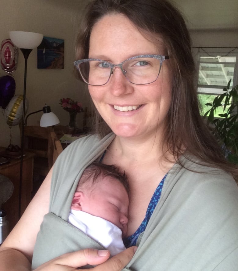 A smiling woman with glasses and long brown hair cuddles a newborn held in a carrier on her chest.