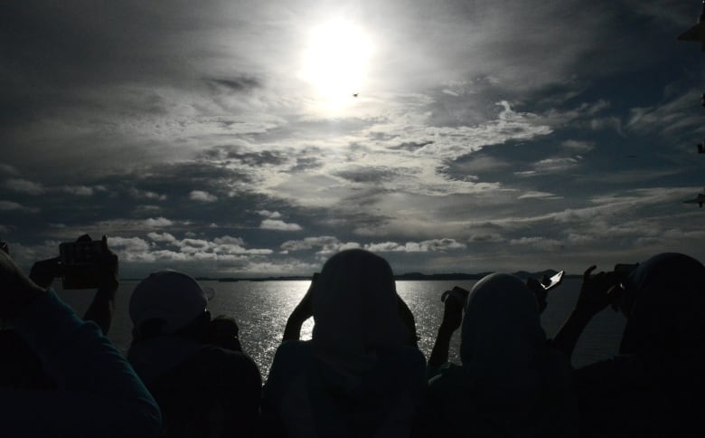 A moody photo shows people from behind gazing out over an ocean with the glow of sun shining through cloudy skies. 