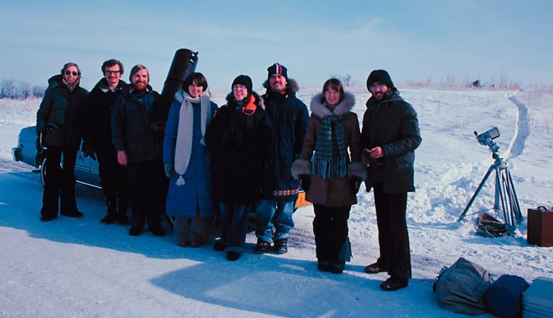 Eight people wearing winter outwear, stand beside a car parked on the shoulder of a highway beside a snow-covered field. Suitcases and gear including a telescope on a stand are nearby. 