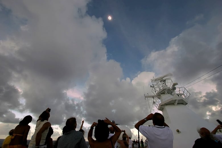 A low-angle photo shows people looking up into the sky where the sun with a black hole in the middle is peeking between the clouds. 