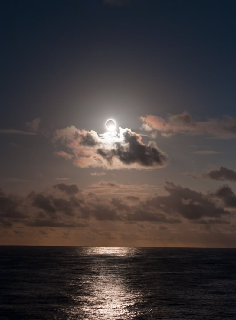 The sun with its centre blocked by the shadow of the moon hangs over the ocean with beautiful clouds in the sky. 