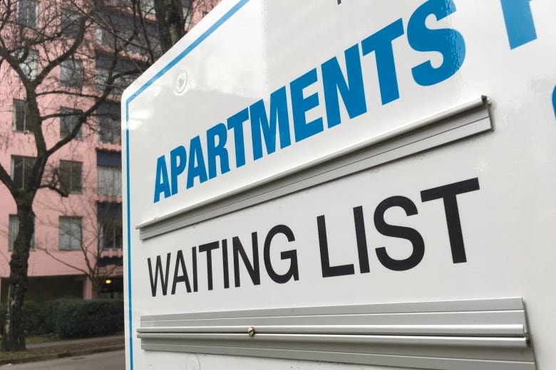 A no vacancy sign with the word Apartments in blue and the words Waiting List in black on a white board in front of a pink brick highrise building.