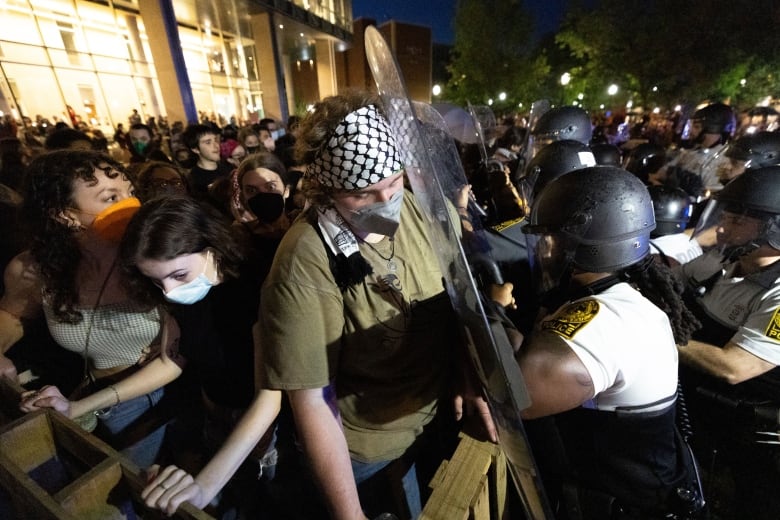 A young man with a headscarf and face covering is pressed up against a riot shield held by a police officer in a nighttime photo.