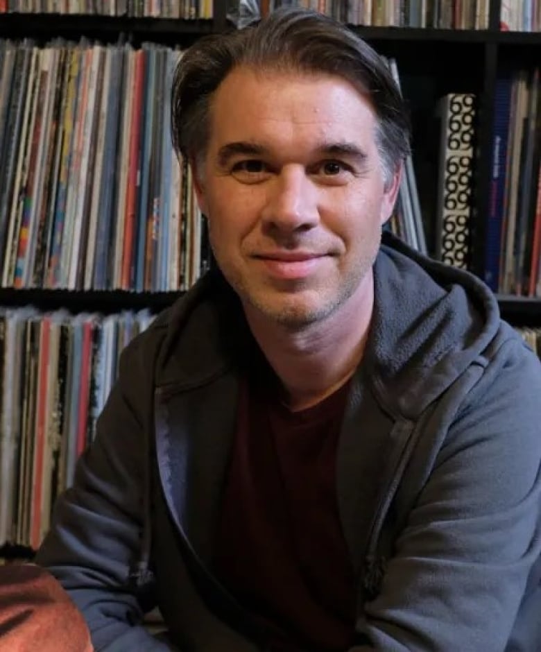 A man is seen smiling at the camera in a close-up shot. He's dressed in a dark grey hoodie.