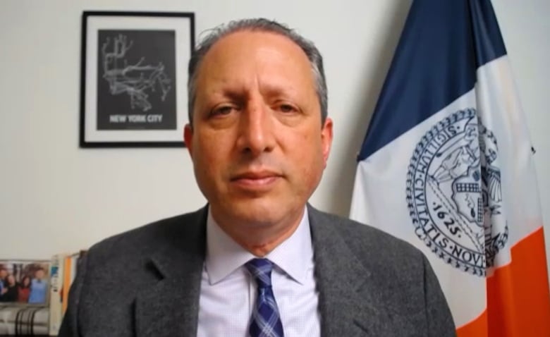 A politician in a grey suit and blue tie sits facing the camera, with the flag of New York City behind him.