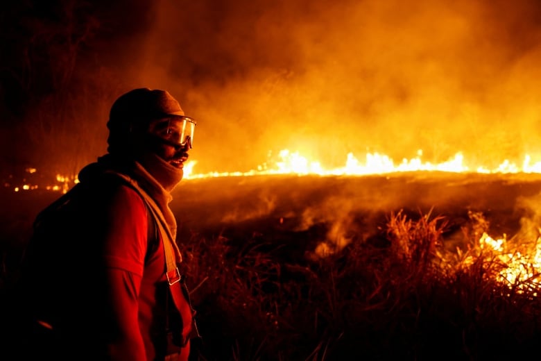 A man in a red suit and goggles stands near a raging fire.