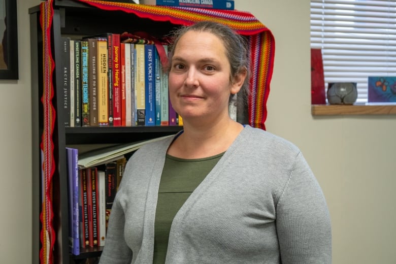 A person wearing a grey sweter over an olive-green shirt stands in front of a bookshelf.