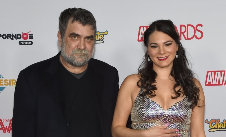 An older man wearing a dark suit jacket and sweater with a grey beard poses with a young dark-haired woman wearing a long, silver dress. 