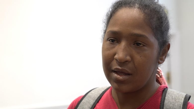 A woman speaks to the camera wearing a pink shirt and grey backpack.