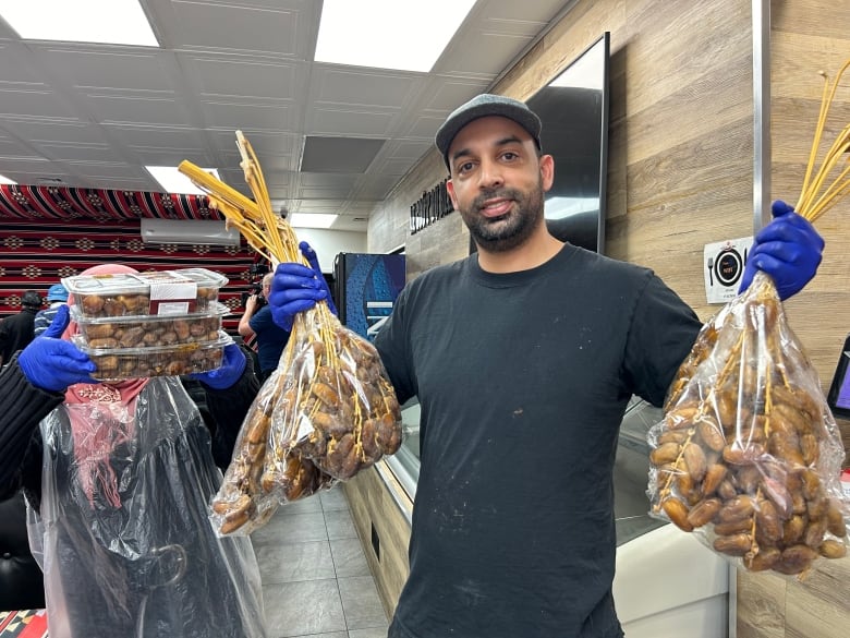 Smiling man holds up two large bags of yellowish dates still on their stems.