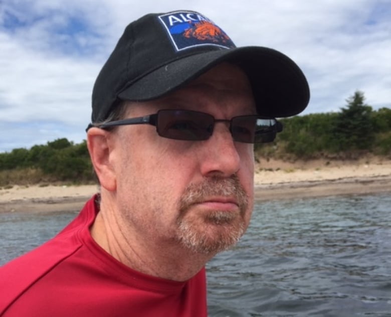 A man with a beard and a baseball cap is seen on the water with the shore in the background.