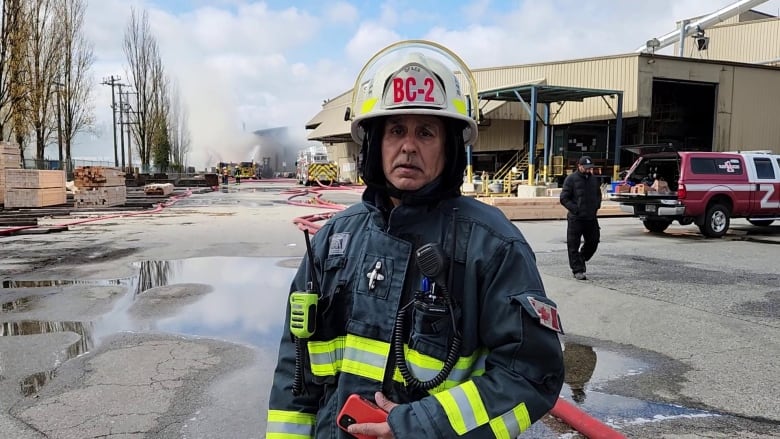 A firefighter looks at a camera while a mill looms behind him in the background.