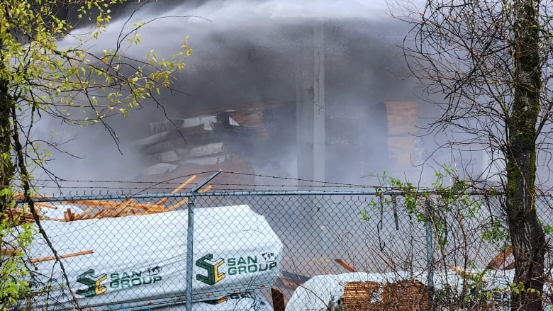 A large cloud of smoke obscures a pile of burned lumber at a factory setting.