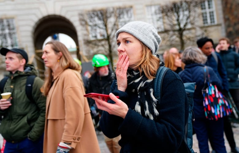 A woman in a grey wool hat stands in a crowd of onlookers and holds her hand to her mouth.