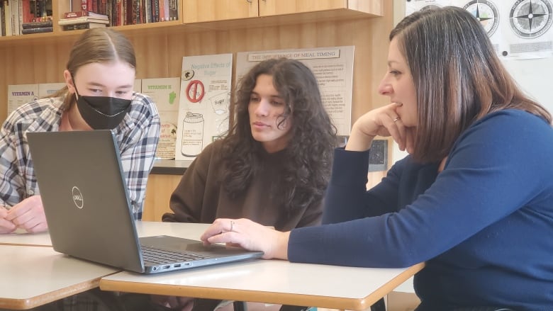 In a bright high school classroom, a teacher scrolls through an open laptop while two students sitting next to her peer at the screen.