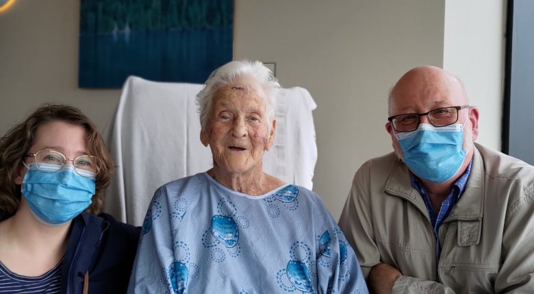 A smiling woman in a hospital gown is seated between a woman and a man who are wearing surgical masks. 