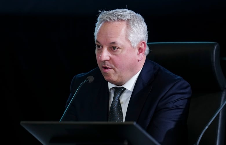 A man in a suit sits at a desk behind a microphone.