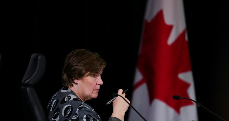 A woman sits in a chair holding a pen. A Canadian flag is drapped in the background.