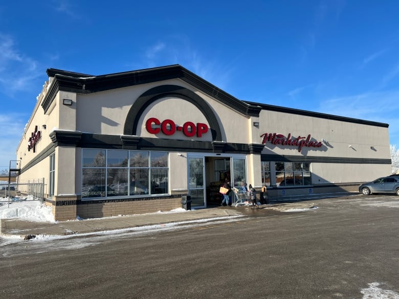 In the foreground is a large parking lot with a but of snow around the edges and one parked car on the right of the image. In the background is a beige and brown building with large windows and automatic sliding doors in the front with one shopper exiting the doors. On the upper left front of the store exterior there is a mounted sign that says "Co Op" and on the right upper exterior there's a mounted sign that says "Marketplace". The sky is almost completely blue above the store.