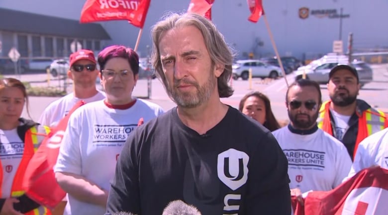 A man stand in front of a group of flag-waving union advocates at a warehouse.